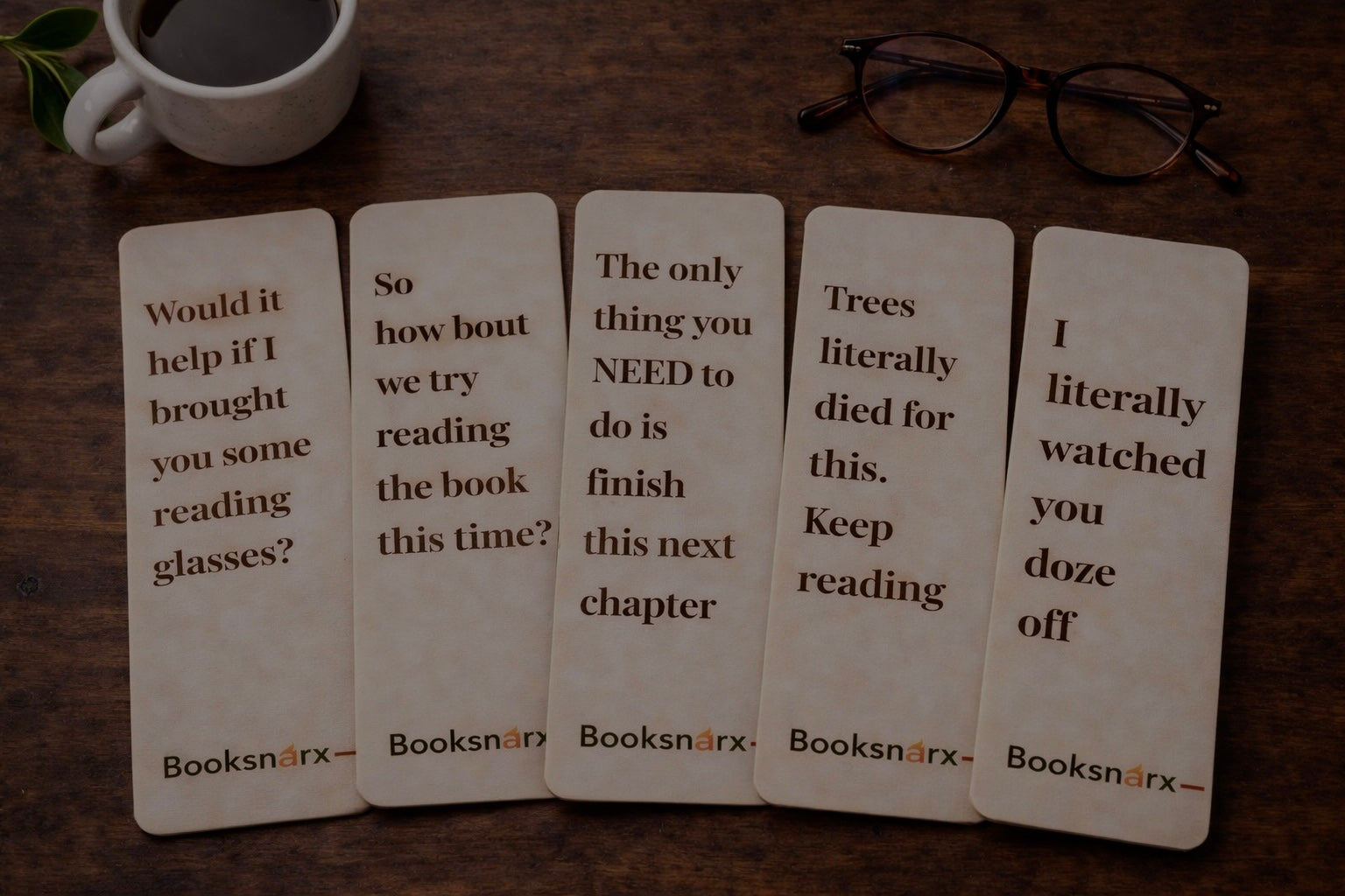 Five bookmarks with humorous text on a wooden surface with a cup and glasses.