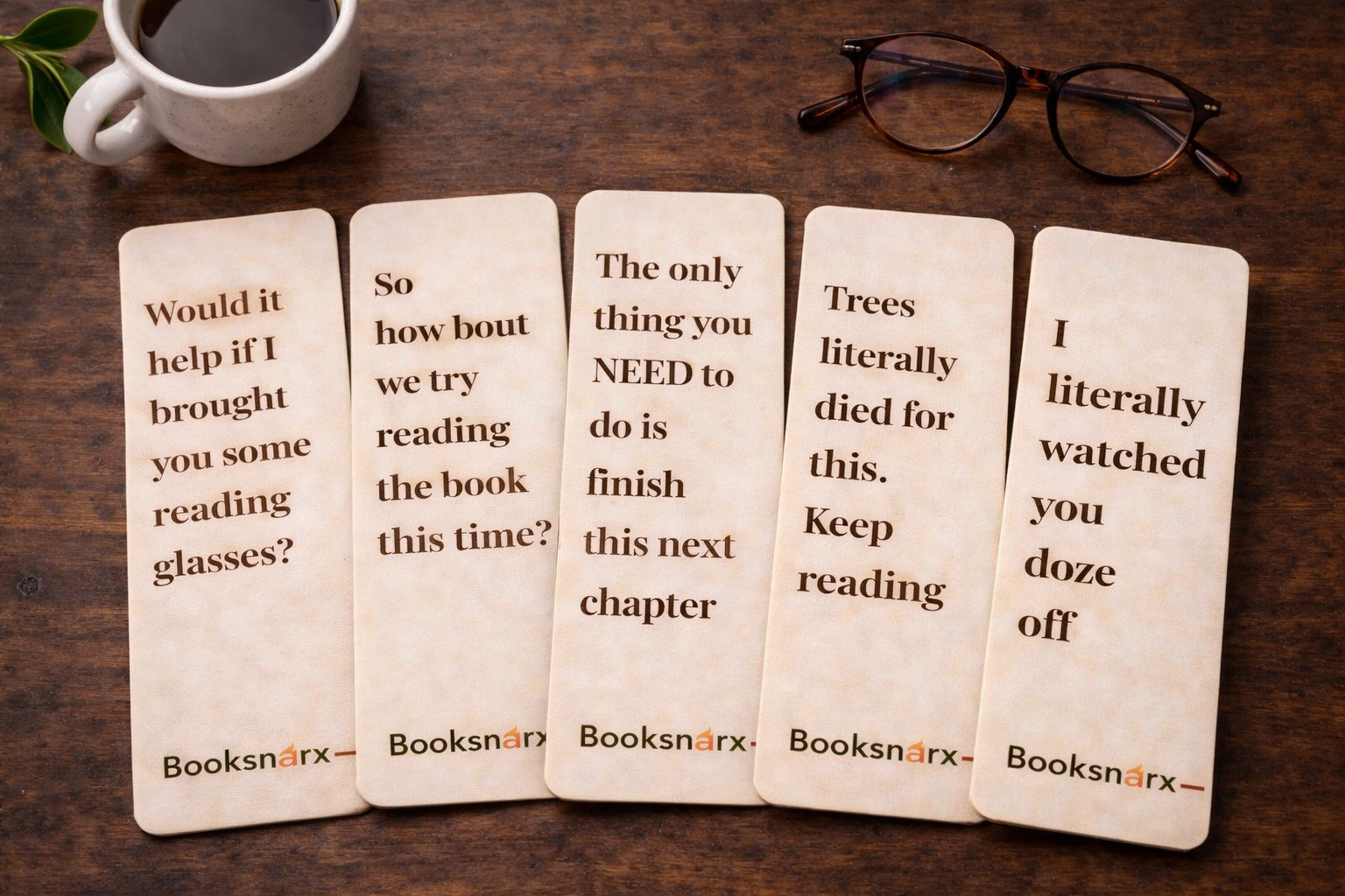 Five wooden bookmarks with humorous text on a wooden surface with a cup of coffee and glasses.