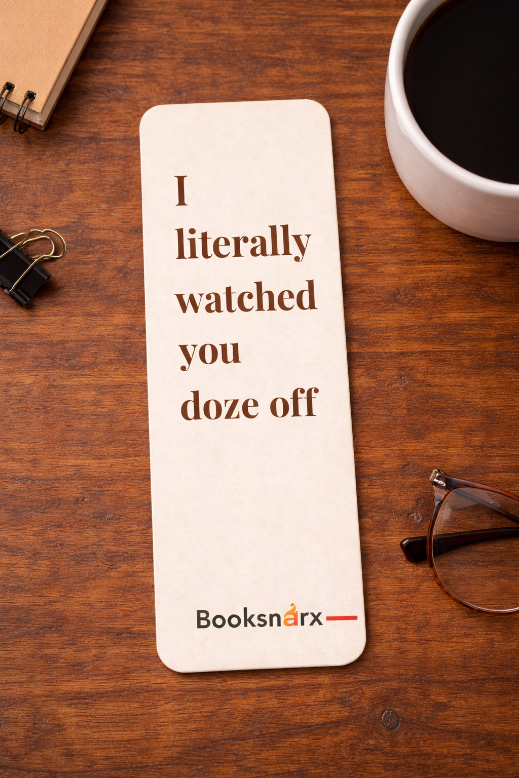 Bookmark with humorous text on a wooden surface with a cup of coffee and glasses.